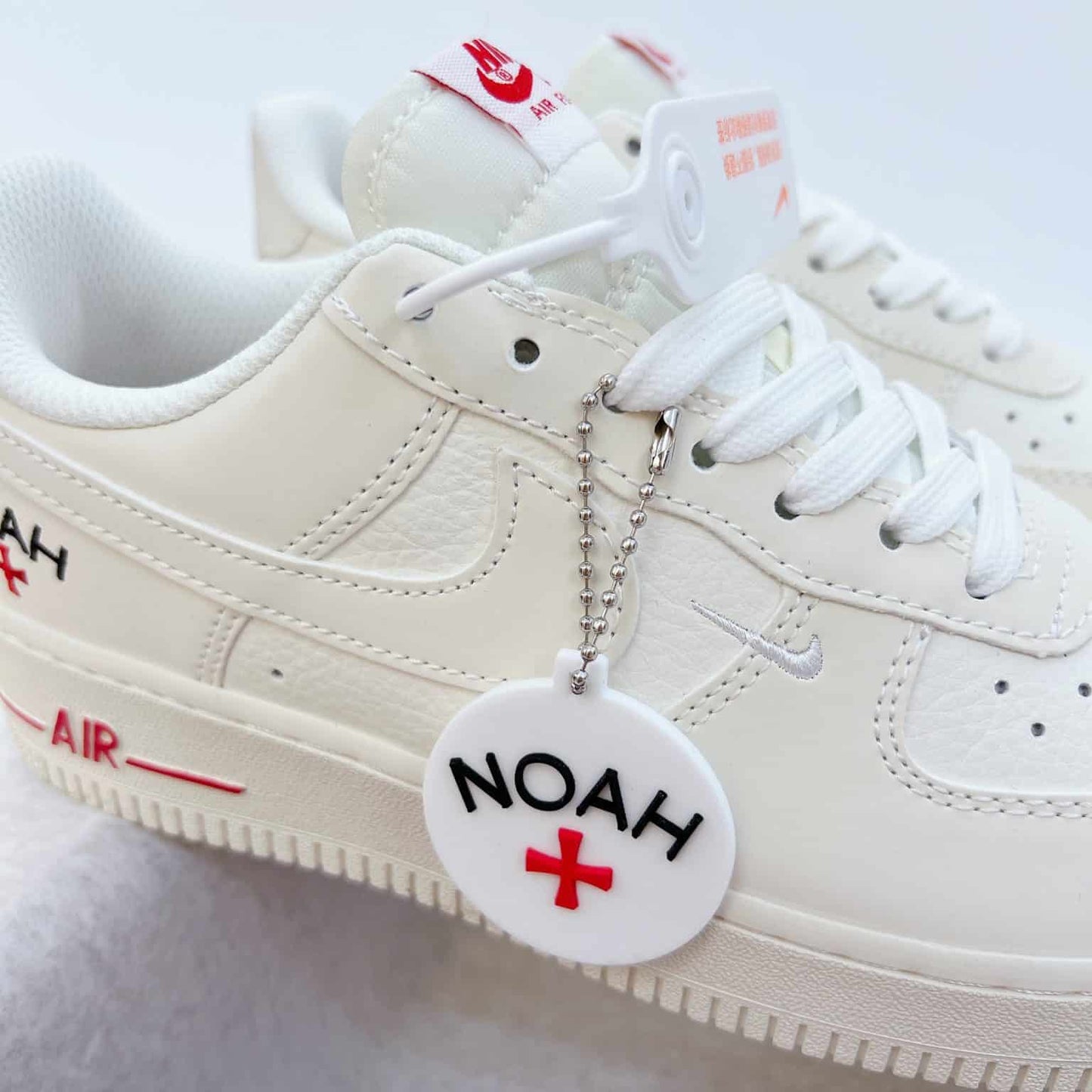 These cream-white Nike Air Force 1 sneakers feature red "Noah" branding accents and a matching circular logo keychain